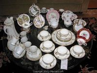 Full set of assorted tea cups and saucers arranged on glass table, showcasing multiple brands and styles with various patterns and colors
