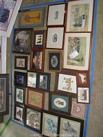 View of various framed country-themed folk art prints arranged on floor including animals, people, and rustic scenes in brown and black frames.