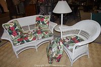 Wicker love seat and chair with floral cushions, table lamp between them, beige woven rug underneath