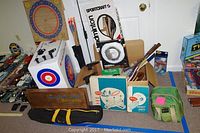 Wide shot showing various sports equipment including dartboards, arrows, rackets, ring toss game, and horseshoes.