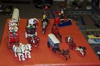 Multiple cast iron reproduction toy wagons and figures displayed on red surface including circus cart, fruit wagon, covered wagons, and horse figurines.