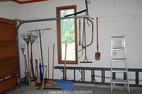 Photo showing rakes, axes, shovels, snow shovels, and 5-foot step ladder in corner of garage