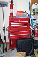 Full view of two stacked red Ace Hardware toolboxes, a fire extinguisher on the smaller top box, and a black cushioned chair back leaning nearby.