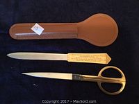 Photo of gold-plated scissors, engraved metal letter opener, and brown stitched leather case on dark fabric background.