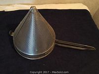 Metal conical perforated kitchen strainer with long handle shown on dark fabric background.