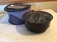 Photo of small portable round black metal BBQ grill next to cushioned insulated carrying case, placed on a bed.