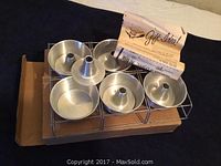 Set of six metal mini ring pans with removable centers displayed on a metal rack, atop a cardboard box with a folded paper insert labeled 'Gift Idea!'.