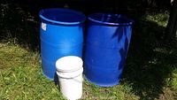 Two large blue barrels and one white 5-gallon bucket outdoors on grass in sunlight.