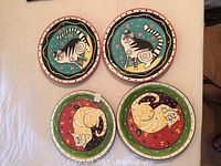 Top down photo showing set of four colorful round ceramic plates hand-painted with whimsical cat motifs in blue, red, green, yellow, black and white.