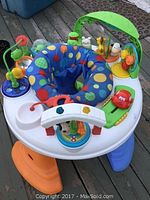 Photo shows a colorful baby exersaucer with a variety of interactive toys attached to a round white base with multicolored legs. The fabric seat is blue with colorful dots. The exersaucer is set on a wooden deck.