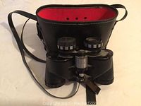 Front view of vintage Carl Wetzlar binoculars with focus adjustment knobs and eyepieces visible, resting in front of a black carrying case with red interior lining and shoulder strap.