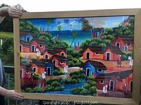 Full view of framed colorful Caribbean village painting with houses, trees, water, and human figures.