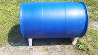Photo of a large blue plastic barrel resting horizontally on a white metal barrel cradle outdoors on grass and gravel.