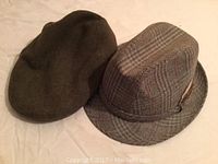 Photo of two hats laid side-by-side on a surface. Left is a dark colored wool newsboy cap and right is a plaid wool blend fedora with feather.