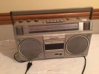 Front view of a silver Panasonic retro portable radio and cassette player, showing dual speakers, cassette deck, analog tuning scale, control knobs, and wooden handle.