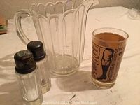 Clear Depression glass pitcher with scalloped edges, two glass salt and pepper shakers with metal tops, and a vintage printed advertisement glass, all shown on a white table.