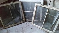 Three antique wooden window sashes on a concrete floor against a wall. Two sashes have two vertical glass panes and one sash with a single large glass pane.