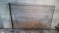 Full view of a metal Edwardian garden fence panel with chain-link mesh and decorative border at top, placed against a concrete wall.