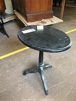 Black round wooden table with single pedestal and tripod leg base, showing minor chips and scratches on surface.