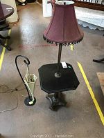 Photo showing both lamps, tabletop lamp on carved wooden pedestal table, floor lamp with attached small table, all on concrete floor.
