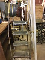 Front view of the metal ladder showing four steps and rusty frame with wheels at base, placed in an indoor area near furniture.
