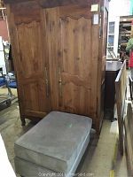 Wooden cherry wardrobe standing next to a gray upholstered ottoman. Wardrobe has traditional detailed door panels and long handles.