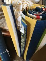 Rolled plastic outdoor rugs showing diverse stripes and patterns in yellow, blue, white, and green tones.