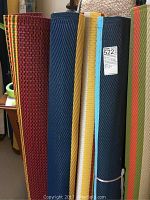 Five rolled plastic rugs in various colors including red, dark blue, light blue, yellow and striped patterns.