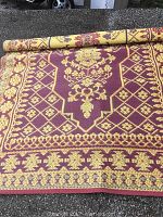 Rolled and unrolled burgundy and yellow patterned plastic outdoor rug on asphalt surface, showing detailed pattern and edges.