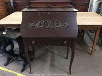 Full view of dark wooden desk with curved legs, fold-down front panel decorated with carved scroll motifs, and one drawer beneath.