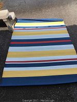 Plastic outdoor rug with bold horizontal stripes in various colors laying unfolded on ground.