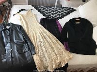 Top view of vintage women's clothing laid out on a bed including a black leather coat, cream lace gown, black wool suit jacket with matching skirt, and black lace shawl.