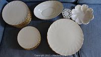 Photo showing stacks of white Lenox China plates with gold scalloped edging, a white oval serving platter, and decorative white serving bowl with gold trim on a blue fabric surface.