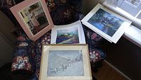 Photo showing four prints arranged on a patterned chair and windowsill, including one framed indoor scene, a colorful water scene, a black and white cityscape sketch, and a rural landscape print.