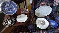 Fifteen collectible plates displayed on chairs and small table, showing a variety of designs including blue and white scenic Wedgwood, black with gold, and floral patterned plates.