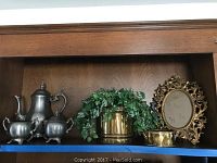 Wide view of shelf showing pewter teapot, sugar bowl, creamer, brass planter with plant, small hammered brass pot, and ornate brass framed mirror.