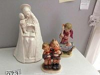 Photo of three figurines: a white glazed Flower Madonna, a pair of boys Hummel figurine, and a Japanese porcelain figurine, shown on a flat surface.