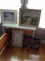 Five framed artworks displayed on a white shelf and the floor near a window showing a signed cottage oil painting, translucent figure art, pencil sketch, and floral canvas.