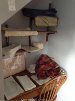 Multiple folded linens including cream/beige placemats, napkins, and a red and black plaid cloth on a chair and wooden shelves