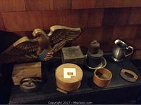 Photo of various vintage decorative items on a shelf including a large carved eagle, small pewter pitcher, old iron, wooden boxes, and kitchen tools.