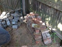 Stacks of used red bricks and gray cobblestones against a wooden fence, outdoors on soil ground.