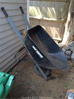 Side view of the metal True Temper wheelbarrow showing the black basin, gray handles with black grips, and single front wheel resting on dirt ground.