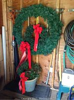 Three green Christmas wreaths with red bow ribbons displayed hanging on wall with garden tools, one large wreath, two smaller wreaths, and a large garden planter below.