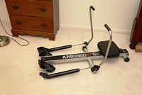 Photo of the full rowing machine showing padded seat, footrests, metal frame, and handles in a room with wooden dresser and lamp.
