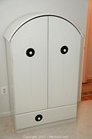 Front view of white Lepine baby armoire with arched top, 2 doors, and bottom drawer showing circular black and white handles.