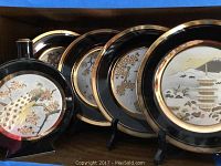 Photo showing four black decorative plates and one decanter, each with gold trimmed edges and detailed Japanese landscape etchings in gold, silver, and copper colors. Plates are on display stands.