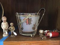 Photo showing glass ice bucket with metal handles and leaf design, two figurine corkscrews, and bottle stoppers on a shelf.