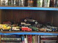 Shelf displays multiple vintage and classic model cars, featuring delivery trucks and sports cars in used condition.