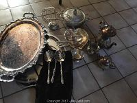 Photo of assortment of silver plated serving pieces including large platters, coffee pot, creamer, sugar bowl, lidded bowl, basket, and serving utensils, all showing tarnish and age wear.