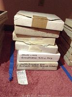 Stack of six flat boxes containing Rockwell collector plates with handwritten labels including 'Grandfather Tells a Story' and 'Rockwell's - Golden Moments Collection'.